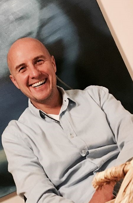 Harald König Harald König an einem Holztisch mit blauem Bild im Hintergrund.
